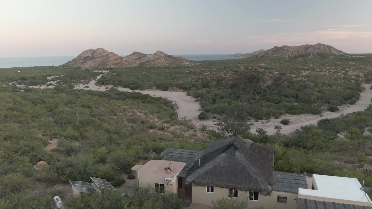 High view of a desert house, solar panels, winding sandy track, and low mountains.
