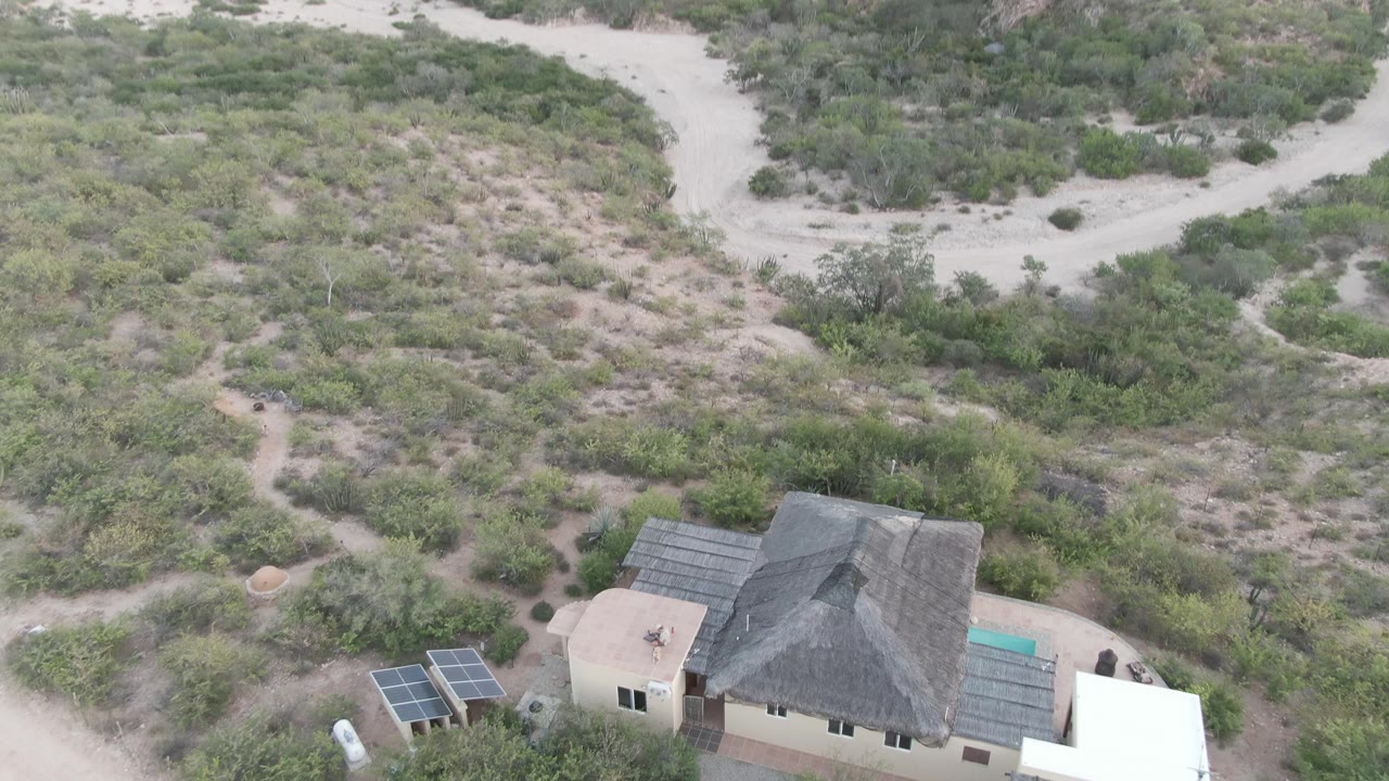 High aerial frame over a Cabo Pulmo desert property, with a dark-roofed house, solar panels, pale dirt tracks, and scrub vegetation, no visible people.
