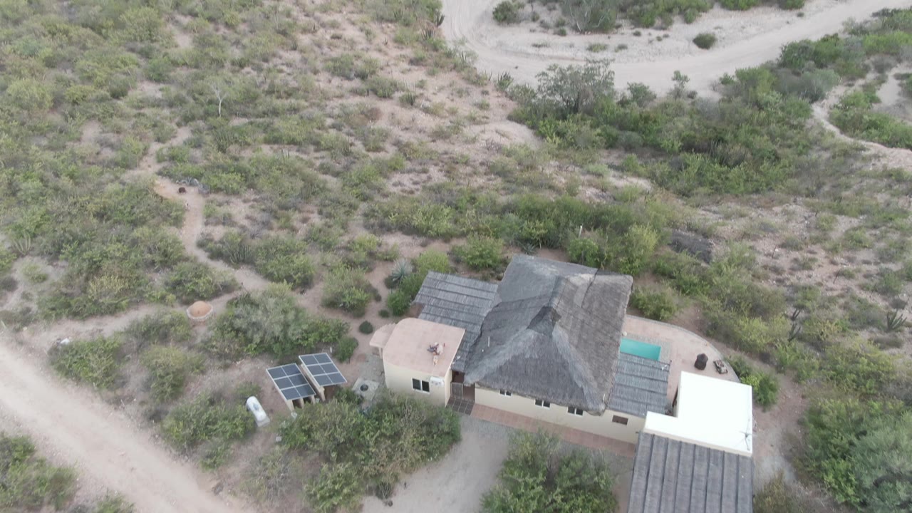 High drone frame over the Cabo Pulmo property, with the dark roof centered low, a turquoise pool edge, solar panels on the left, and pale dirt roads crossing the scrubland, no visible people.
