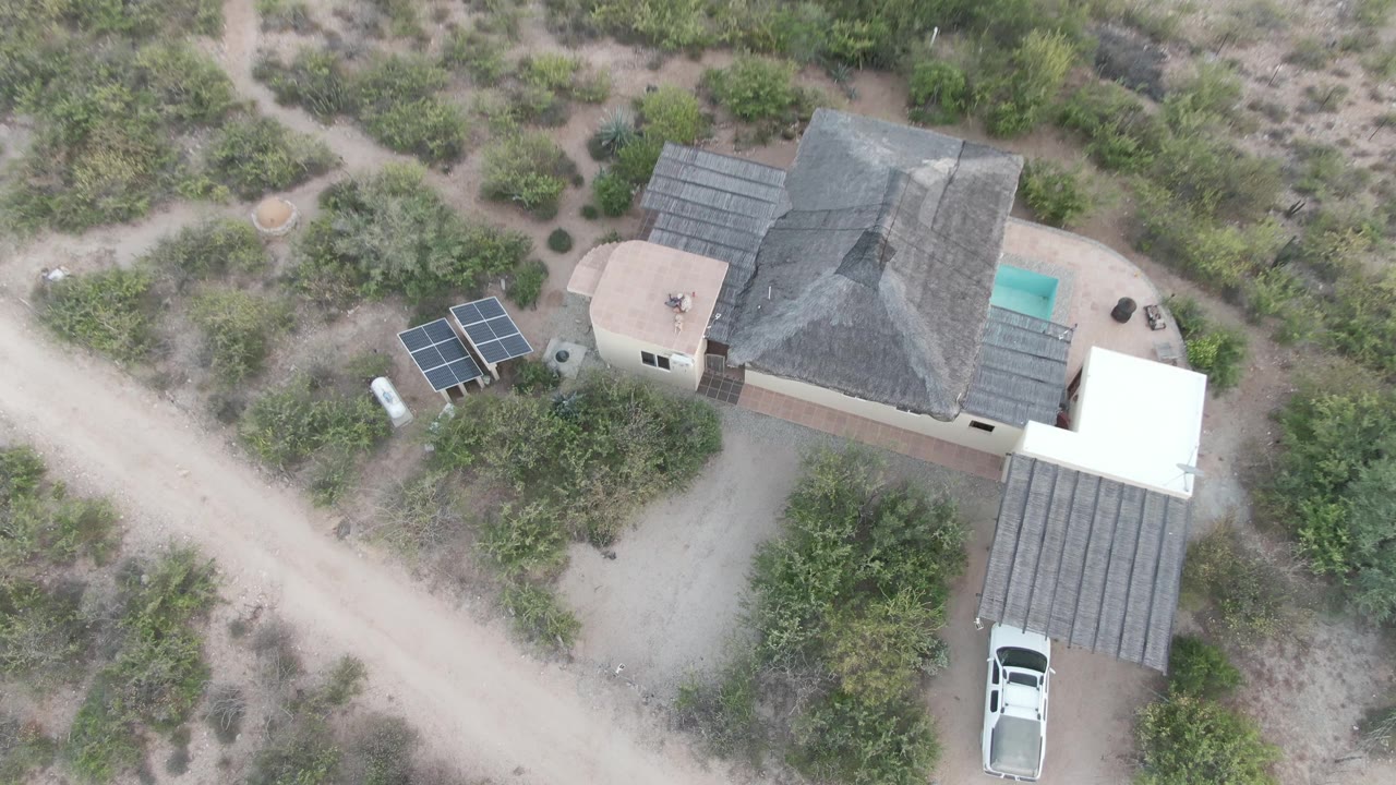 Overhead frame in Cabo Pulmo, Mexico showing a desert home, solar panels, dirt road, sparse bushes, and a white vehicle still mostly under the carport, with no visible people.
