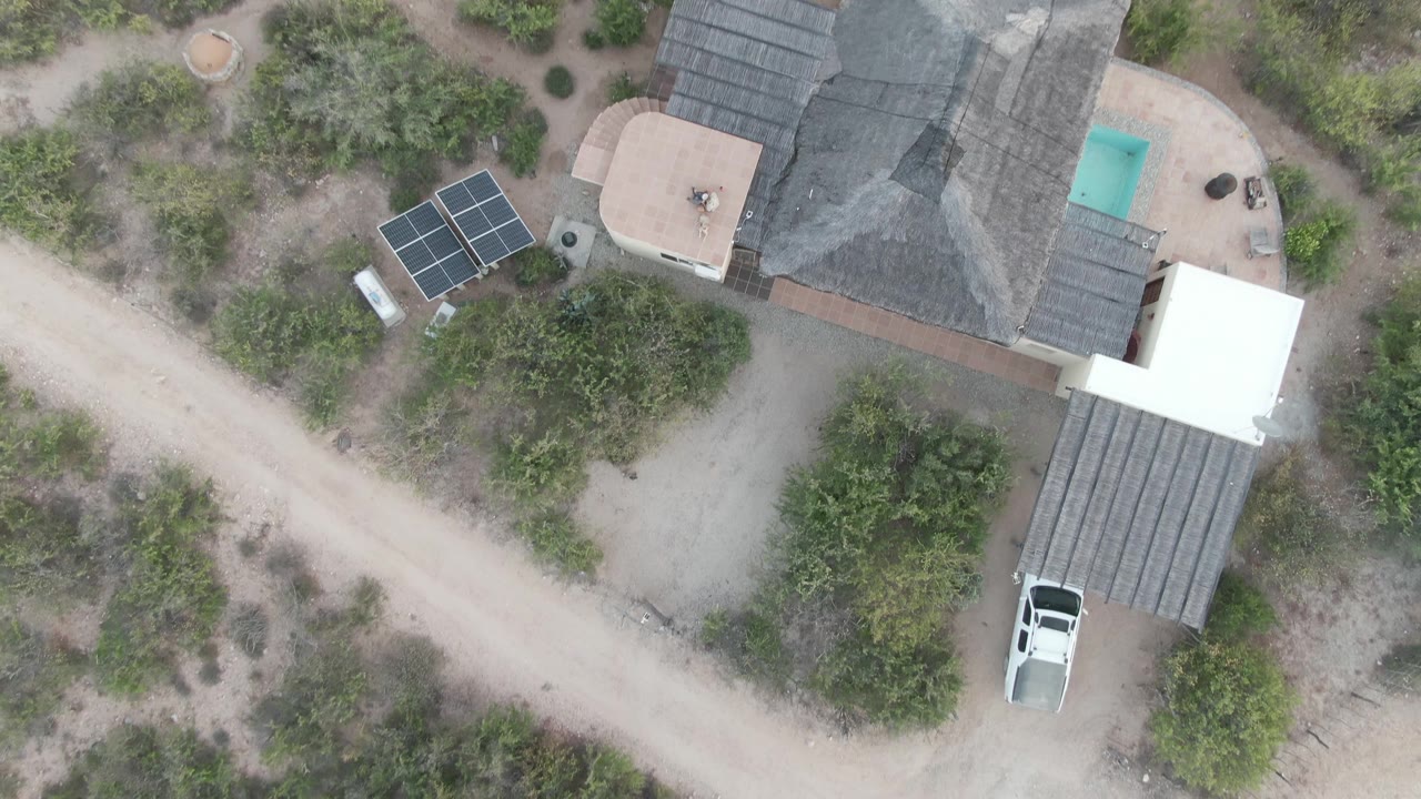 Top-down Cabo Pulmo frame showing the white vehicle farther out from the carport, with the dark-roofed house, solar array, dirt road, patio, pool, and desert shrubs visible and no visible people.