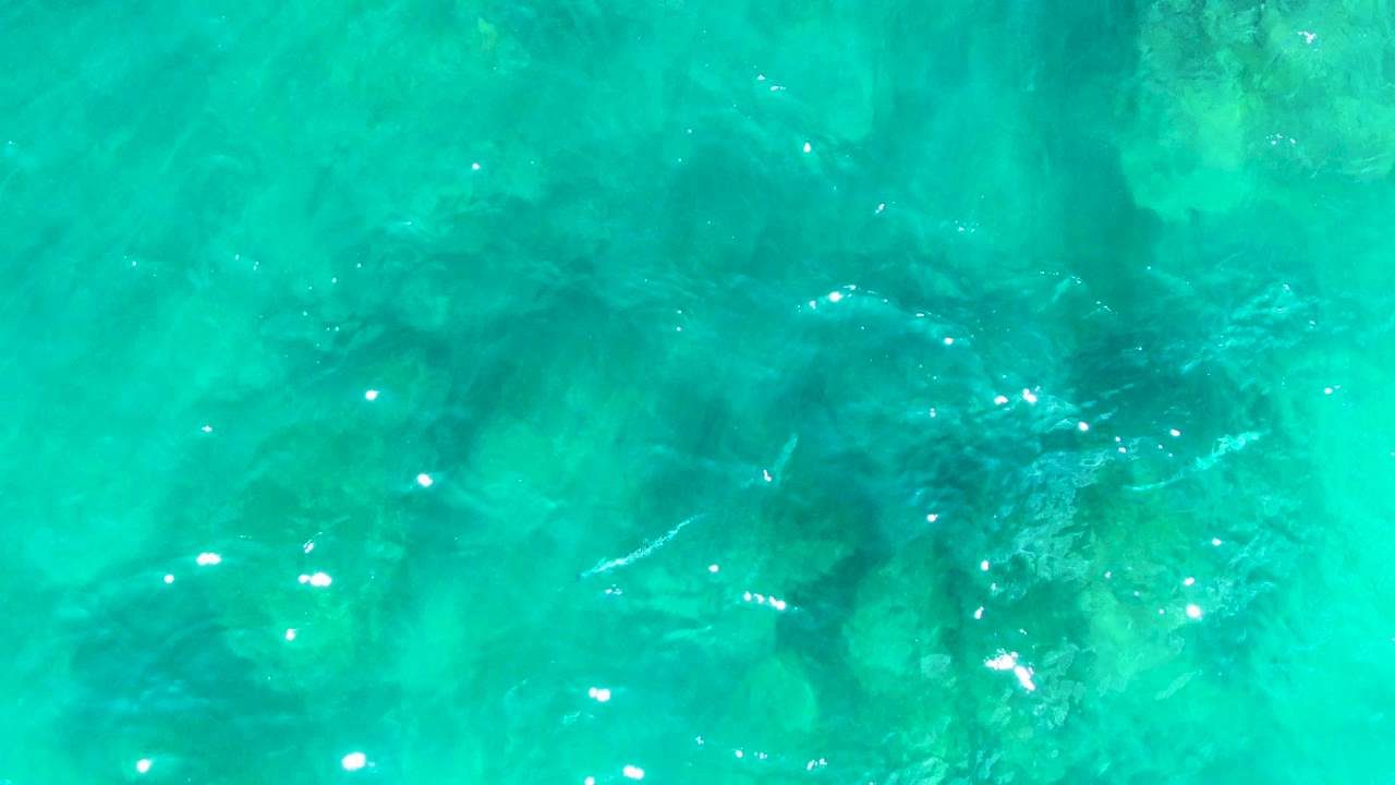 Turquoise water at Punta Buzzards reveals a pale greenish underwater rock patch near the right side, darker rock shapes below, and no visible people.