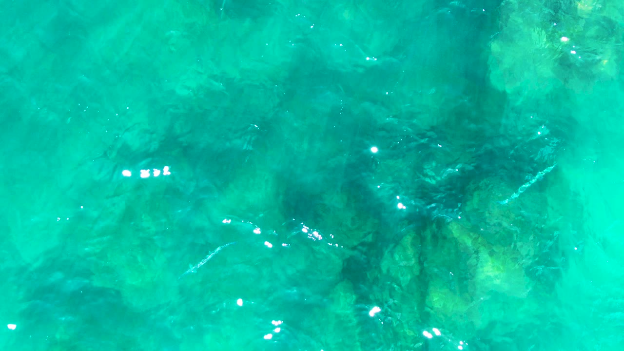 Rippled turquoise ocean at Punta Buzzards covers shadowed underwater rocks, with small white sun reflections and no visible people.