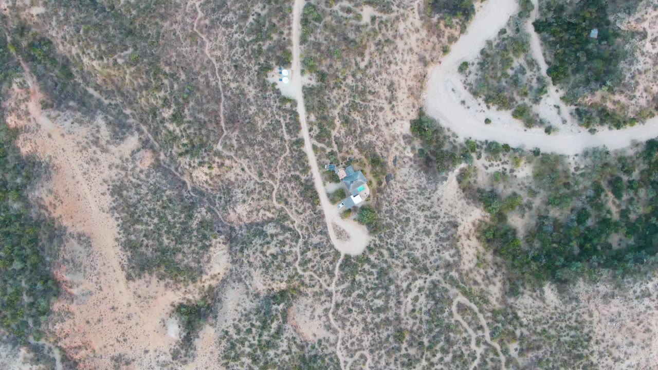 At 0:01, the Cabo Pulmo aerial composition remains nearly unchanged, with a pale road bending around green scrub and the small structure still visible, with no visible people.