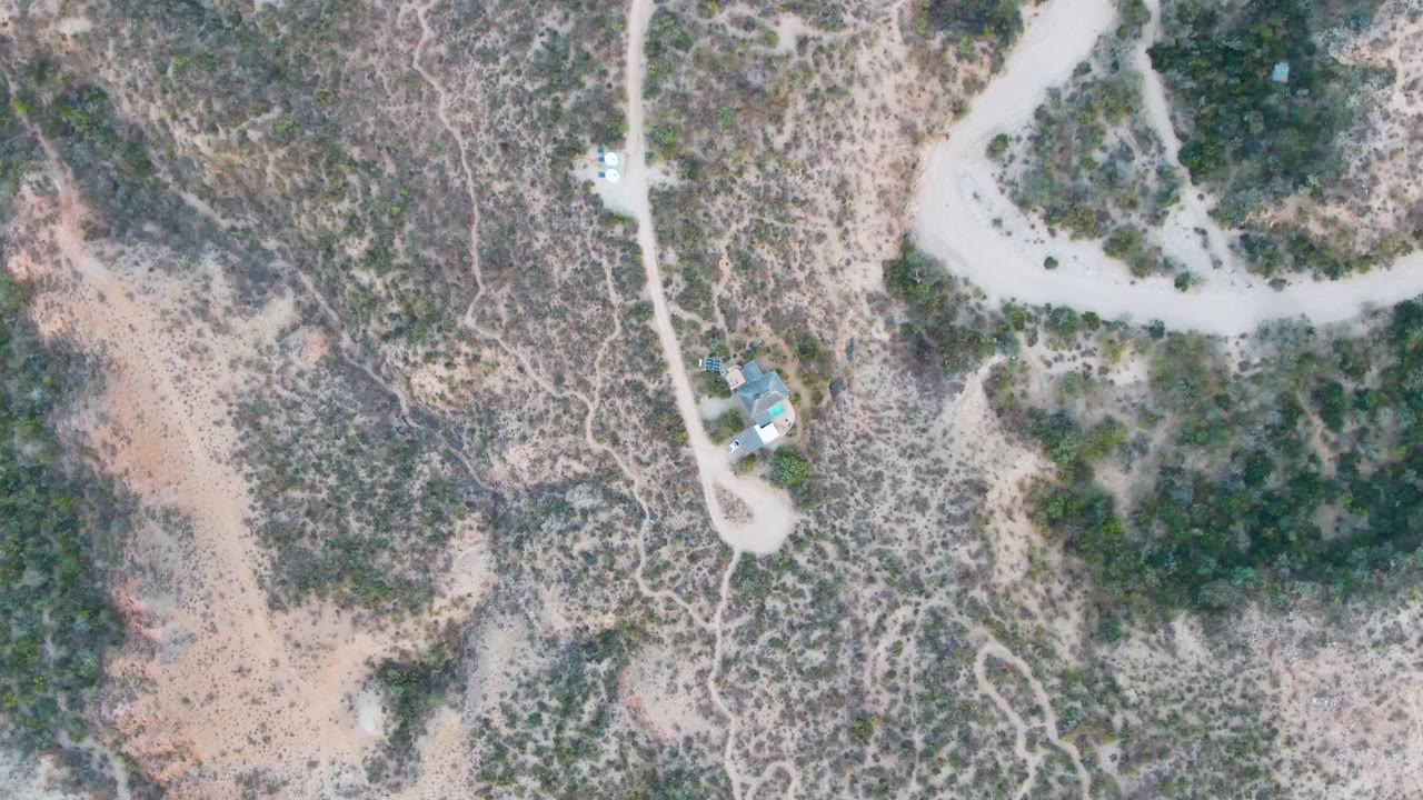 Overhead view in Cabo Pulmo, Baja California Sur with the dirt road curving near small structures, mottled scrubland, rocky soil, and no visible people.
