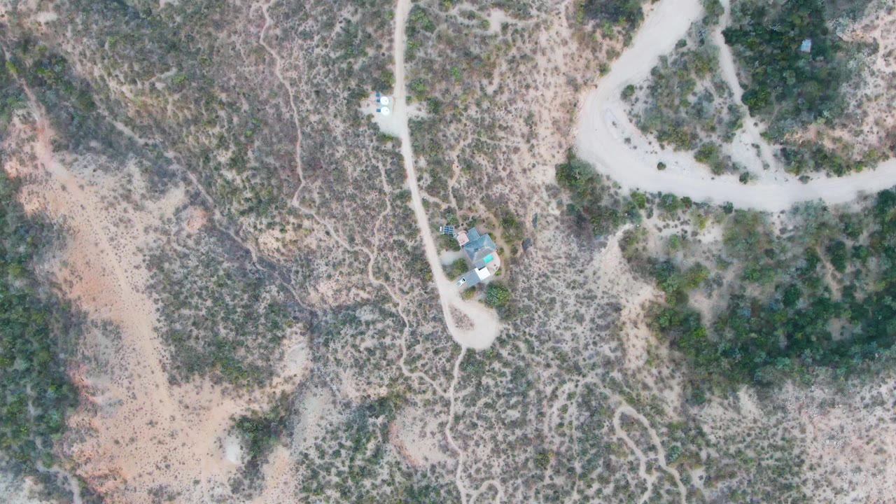 Aerial frame of a pale dirt road beside scrubby desert hillside terrain.