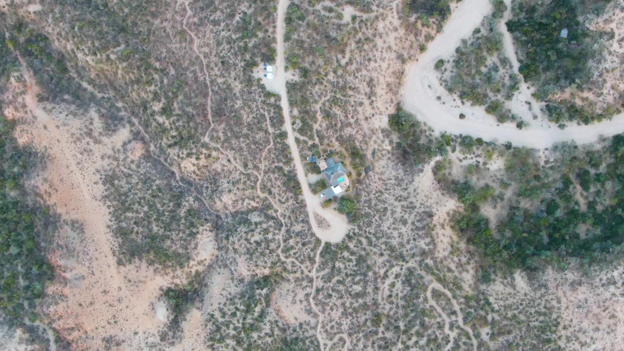 Overhead aerial frame in Cabo Pulmo, Baja California Sur, showing dry scrub terrain, intersecting pale dirt roads, a small light-roofed building near the road, and no visible people.