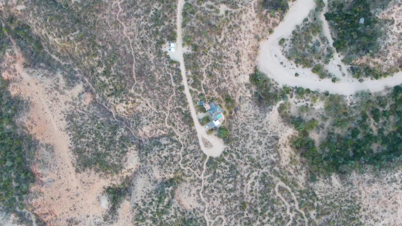 One second in, the static aerial frame keeps the Cabo Pulmo dirt road, scrub-covered ground, and small structures in nearly the same overhead composition with no visible people.
