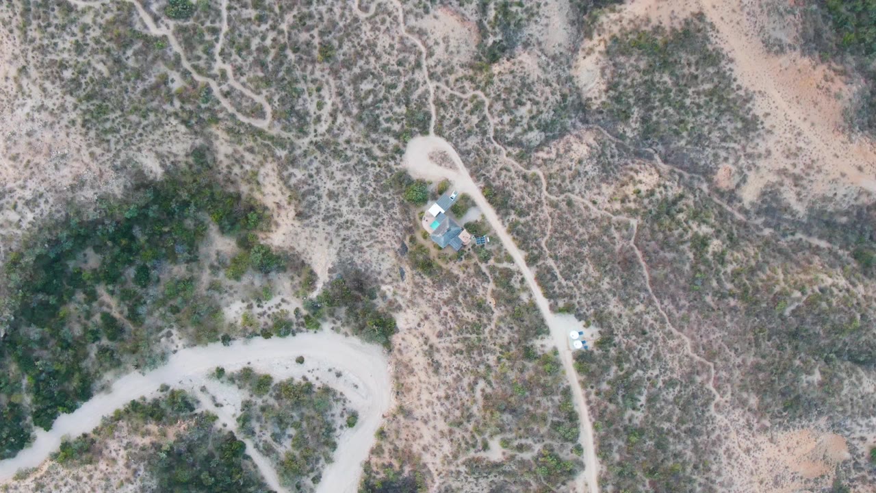Overhead Cabo Pulmo frame showing a light dirt road, a small vehicle on or near the track, a nearby structure area, scrub-covered slopes, and no visible people.