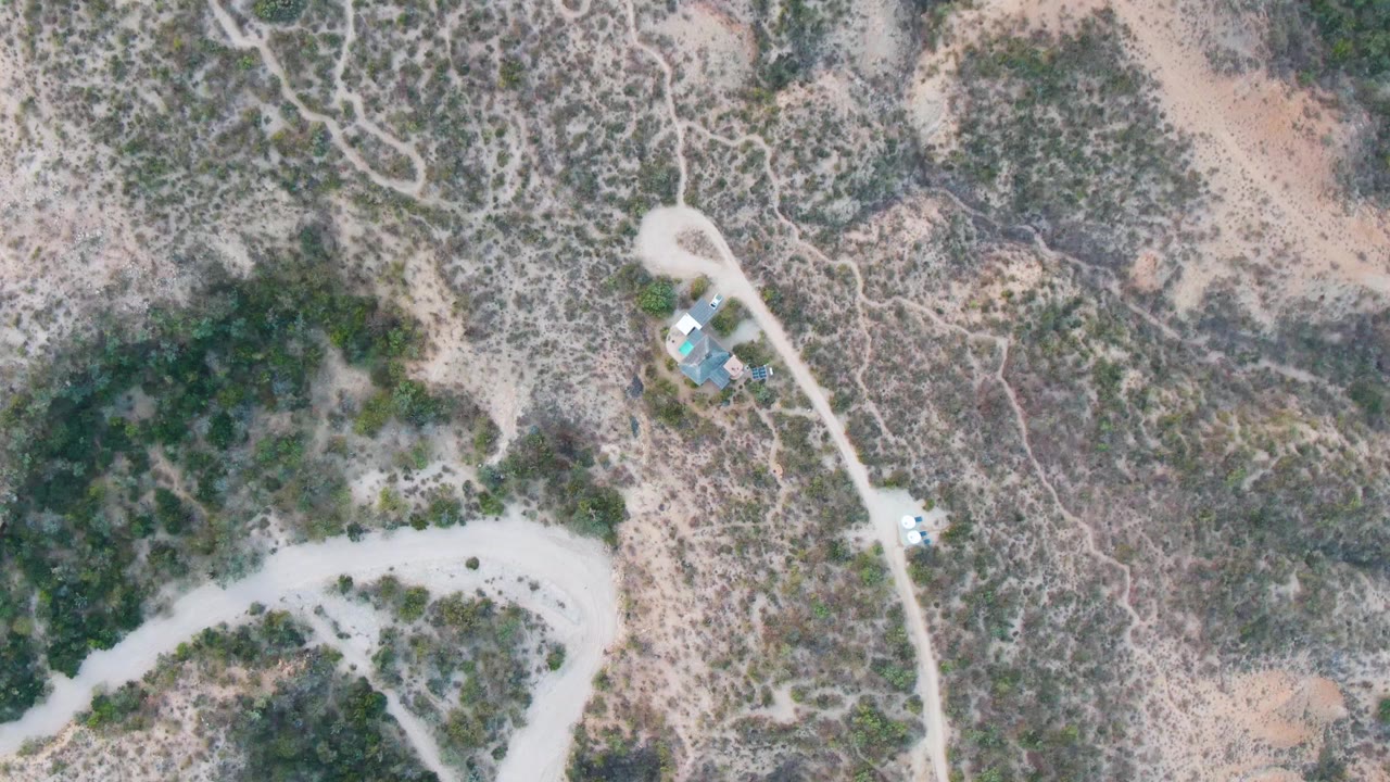Overhead view in Cabo Pulmo, Baja California Sur, showing pale dirt roads, scrubby dry terrain, small blue-roofed structures, a small vehicle near the road, and no visible people.