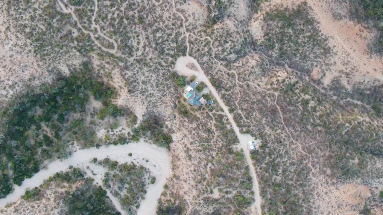Second overhead frame in Cabo Pulmo shows the curved dirt road, dry hillside vegetation, small structures near the upper road, a light vehicle, and no visible people.