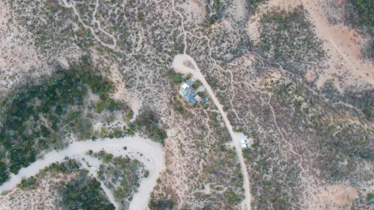 Mid-clip aerial frame in Cabo Pulmo shows tan and gray-green desert terrain crossed by dirt roads, a small vehicle lower on the road, small blue-roofed structures, and no visible people.