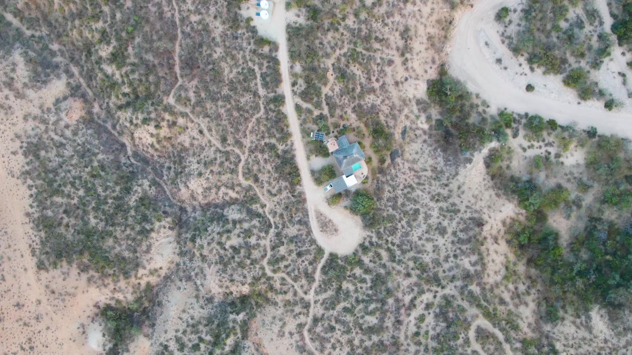 High aerial view over Cabo Pulmo desert terrain with pale dirt roads, scattered shrubs, a small blue-roofed house, a tiny vehicle near the upper road, and no visible people.