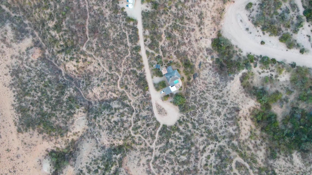 Overhead frame from Cabo Pulmo showing a small house beside a curving dirt road, mottled desert shrubs, sandy ground, a small vehicle on the road area, and no visible people.