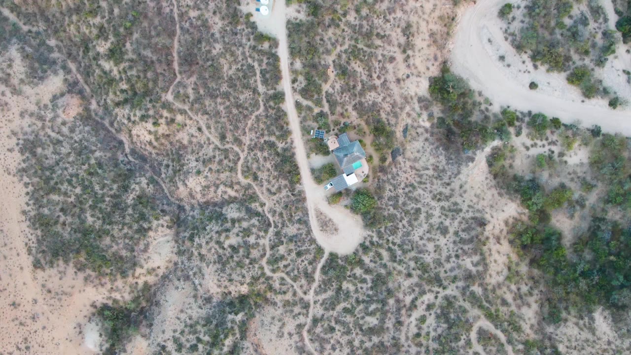 Cabo Pulmo aerial frame with the blue-roofed house near the lower road bend, a pale road running upward, desert brush across the slopes, a tiny vehicle, and no visible people.