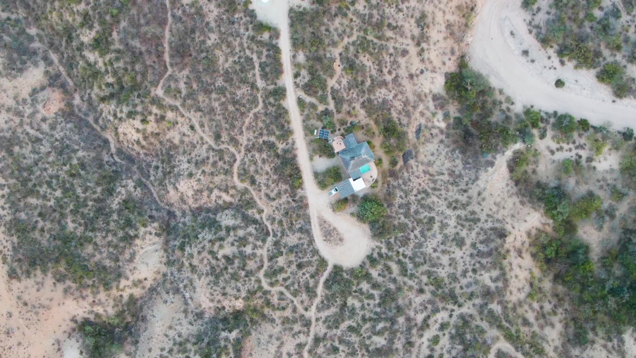 Aerial frame in Cabo Pulmo, Baja California Sur with the small building below center-right, winding pale dirt roads, dry scrubland, and no visible people.