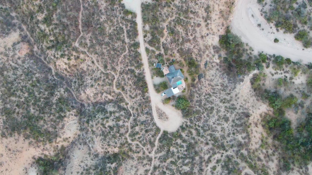 Overhead Cabo Pulmo frame showing the small building near a dirt-road junction, mottled desert vegetation, pale sandy ground, and no visible people.