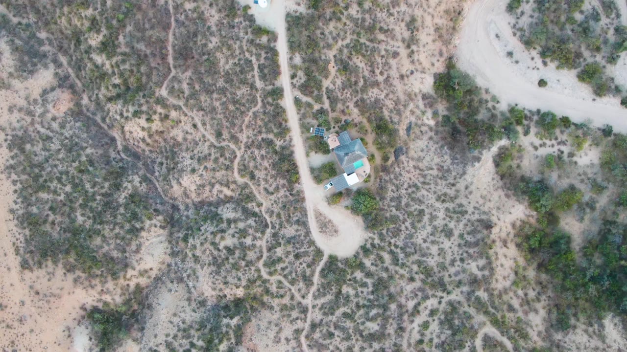 High overhead Cabo Pulmo frame showing an isolated house, winding dirt tracks, patchy shrubs, and pale sandy ground, with no visible people.