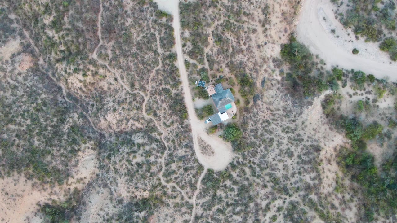 Overhead view in Cabo Pulmo, Baja California Sur of a pale dirt road curving past a small house and vehicle amid sandy desert scrub, with no visible people.