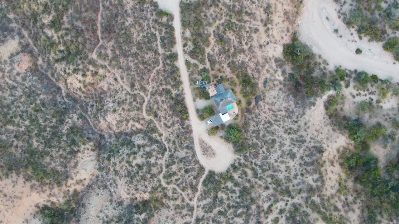 Aerial Cabo Pulmo frame with the car on the light dirt road, the small house set just off the road, and dry scrub spreading across the surrounding terrain, with no visible people.