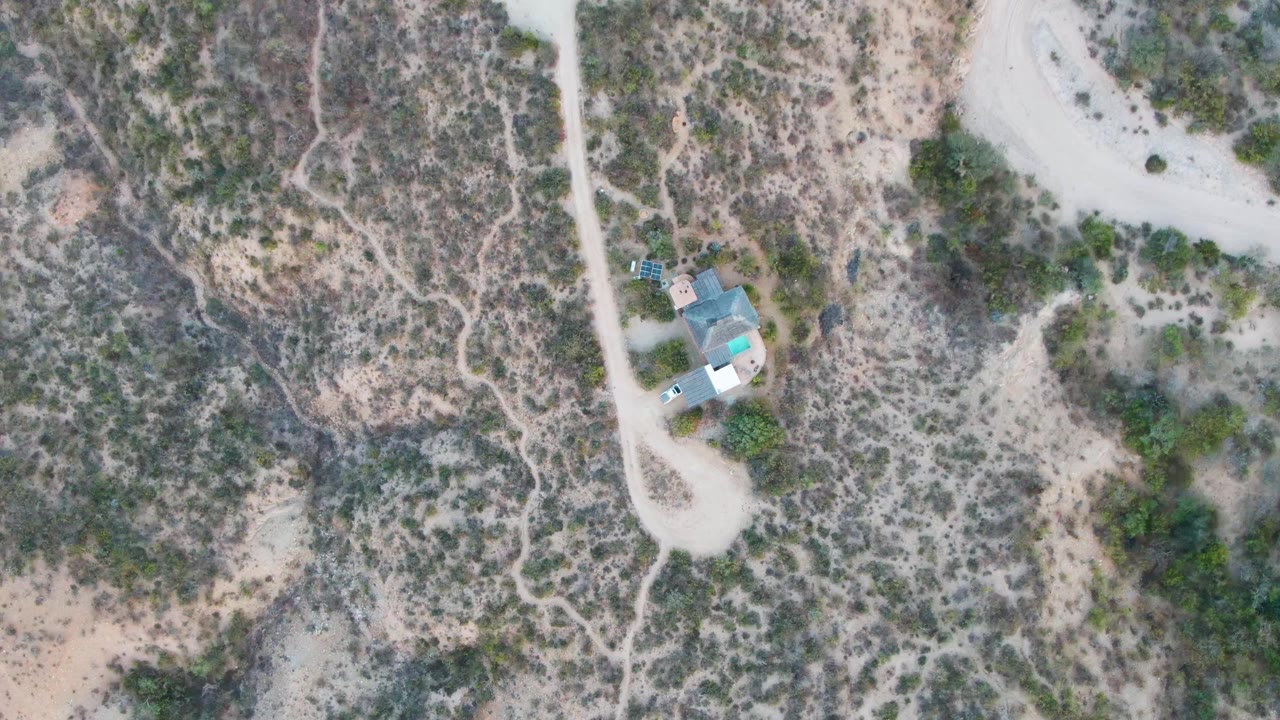 Top-down view over Cabo Pulmo desert terrain, showing the vehicle beside the small house, branching footpaths, pale sand, and scattered vegetation, with no visible people.