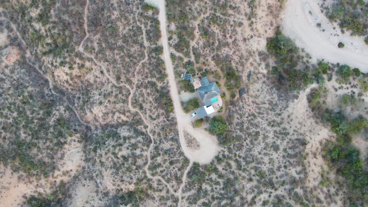 High overhead view in Cabo Pulmo with the pale dirt road running past the small building, scrub vegetation across sandy ground, and no visible people.