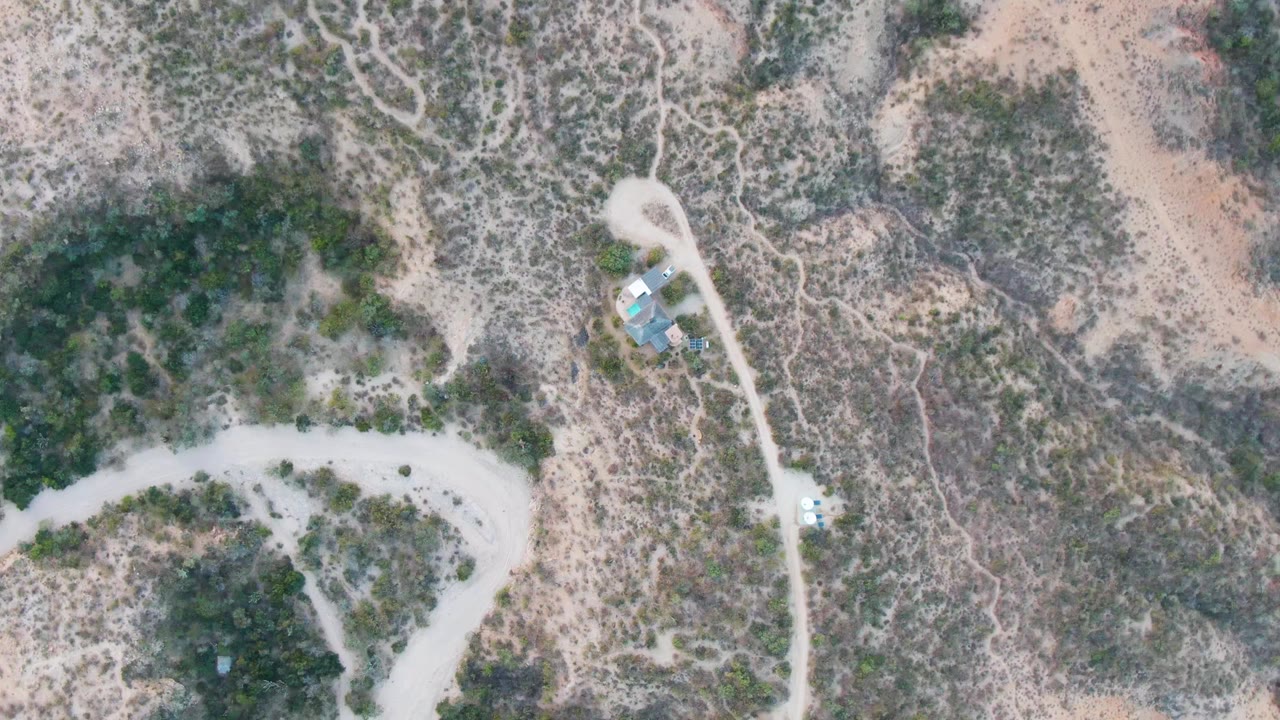 Overhead view in Cabo Pulmo, Baja California Sur, showing a pale dirt road curving through dry scrub with a small blue-roofed structure nearby and no visible people.
