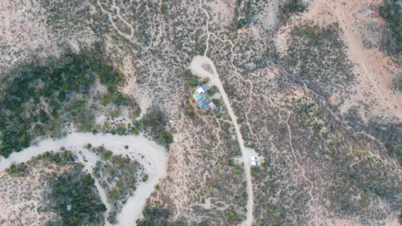 A high aerial frame in Cabo Pulmo shows the road bending across sandy terrain, with scrub-covered patches, faint side tracks, a small structure, and no visible people.