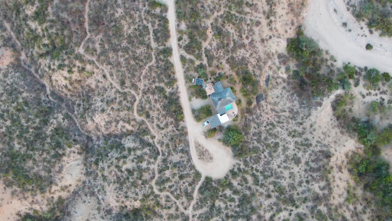 Overhead frame in Cabo Pulmo, Baja California Sur showing a pale dirt road, scrubby arid ground, a small roofed building cluster, and a light vehicle nearby, with no visible people.