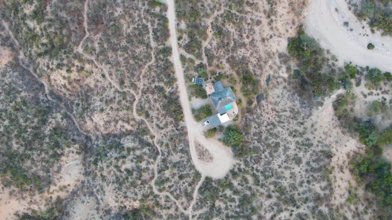 Aerial Cabo Pulmo frame showing the road curving past sparse vegetation and the small building cluster set against mottled tan and green desert ground, with no visible people.