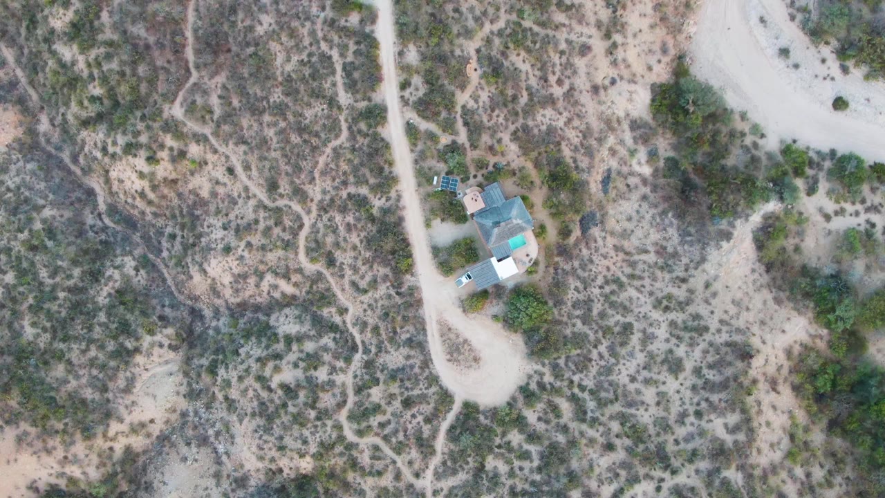 Top-down view in Cabo Pulmo with pale roads crossing dry scrub, the roofed buildings near the lower-left area, and no visible people in the frame.