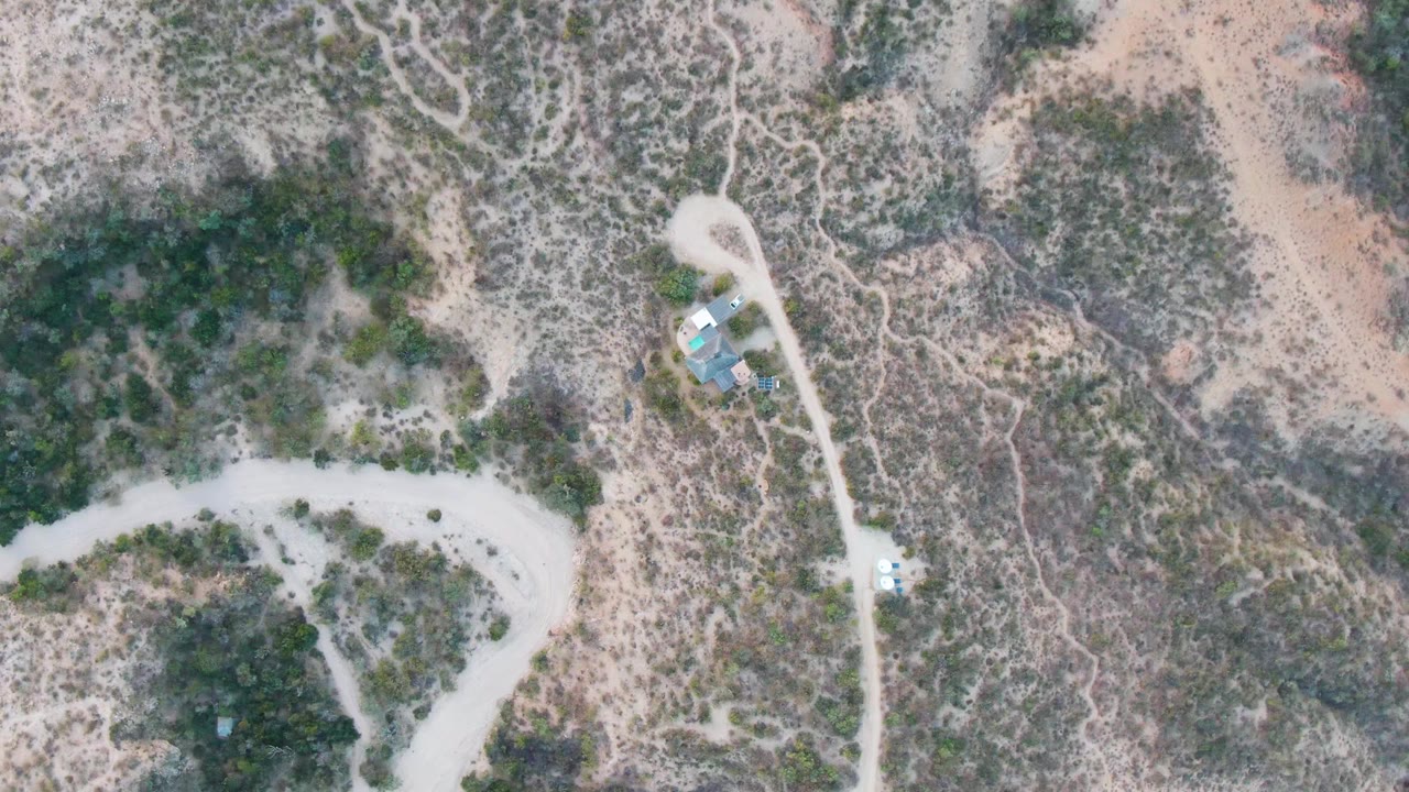 Overhead drone view in Cabo Pulmo, Baja California Sur, showing a pale dirt road, scrub-covered desert, sandy washes, small roofed structures, and no visible people.