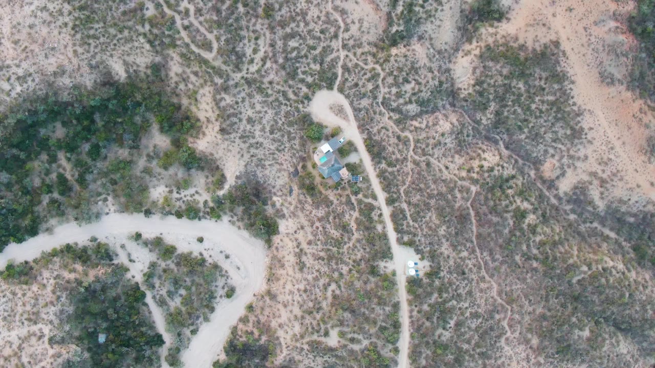 Aerial view over Cabo Pulmo desert terrain with the road running vertically past a small group of structures, scattered shrubs, dry sand, and no visible people.