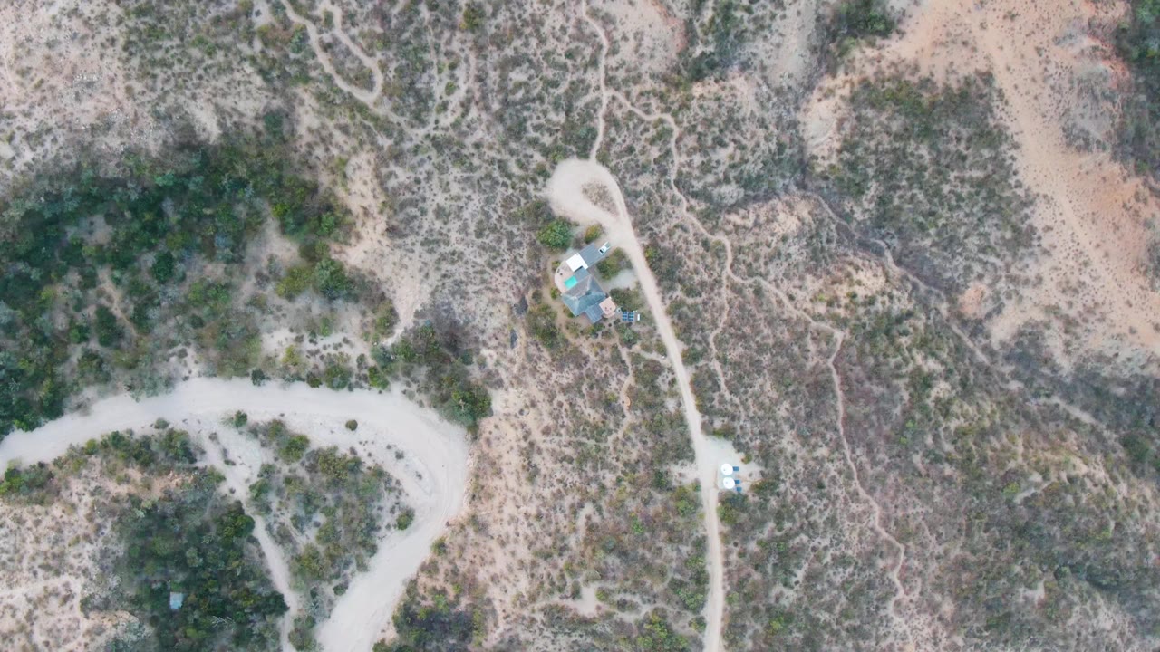 Top-down Cabo Pulmo frame with a light vehicle on the pale dirt road below the roofed structures, surrounded by mottled desert brush and no visible people.