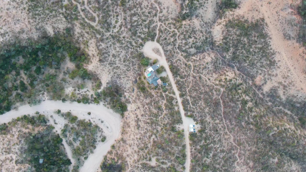 Top-down Cabo Pulmo desert frame with a winding pale dirt road, a small cluster of structures beside the track, scattered shrubs, rocky ground, and no visible people.