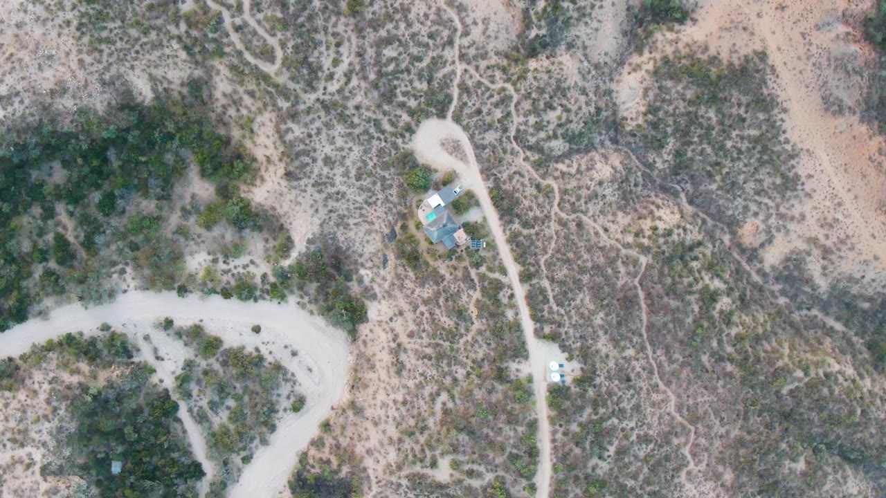 Aerial frame over Cabo Pulmo showing intersecting dirt tracks, mottled tan and gray desert vegetation, small roofed structures near the road, a small vehicle shape, and no visible people.