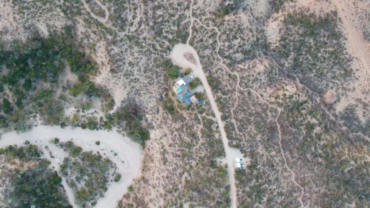 Aerial frame in Cabo Pulmo with the white vehicle slightly farther along the pale dirt road, a compact structure cluster near the road bend, scrub vegetation, sandy ground, and no visible people.