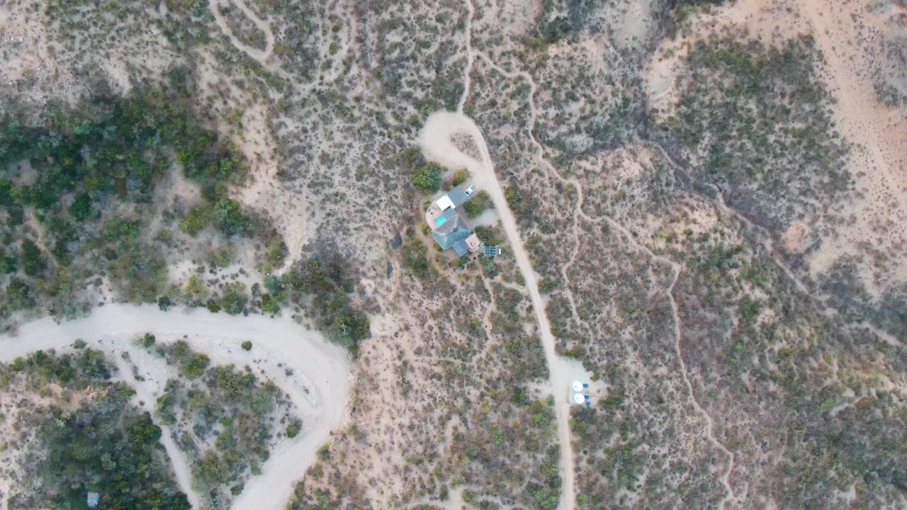 High overhead Cabo Pulmo frame showing the vehicle on a pale dirt road, a cluster of small blue and light roofs near the road, dense scrub patches, dry ground, and no visible people.