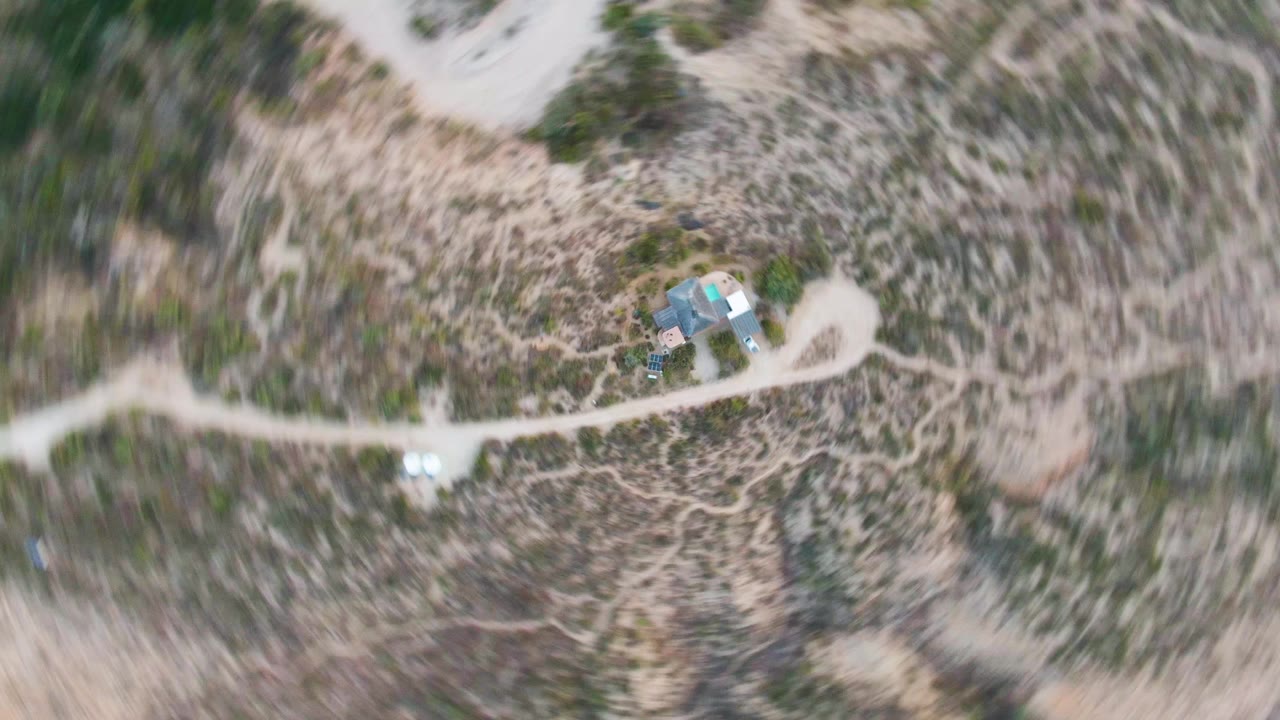 Direct overhead frame in Cabo Pulmo, Baja California Sur, with the cabin closer to the lower edge, a curving dirt track, dry brush, and no visible people.