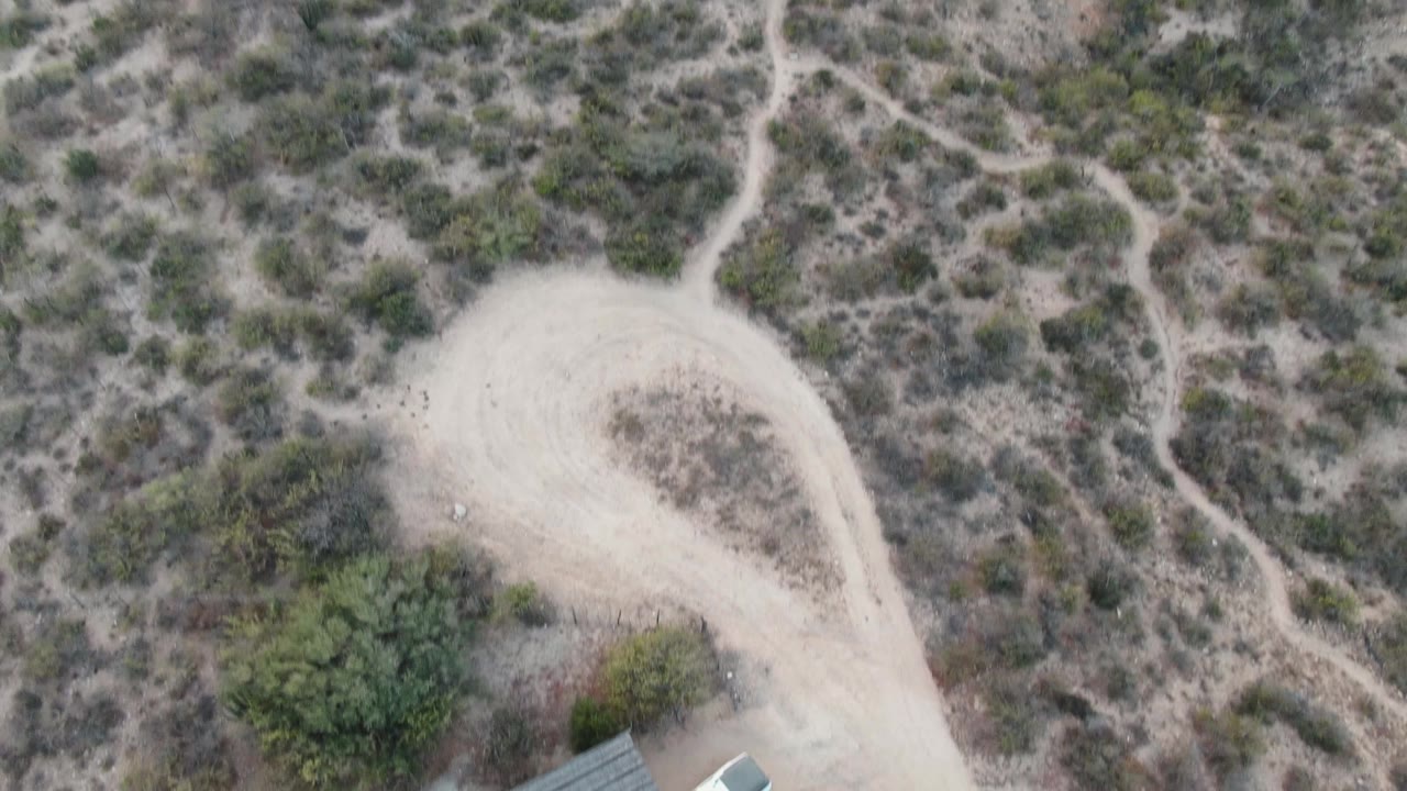 Aerial frame over Cabo Pulmo desert terrain with a curved sandy road, dense scrub patches, and the edge of a small roof beginning to appear at the lower right, no visible people.