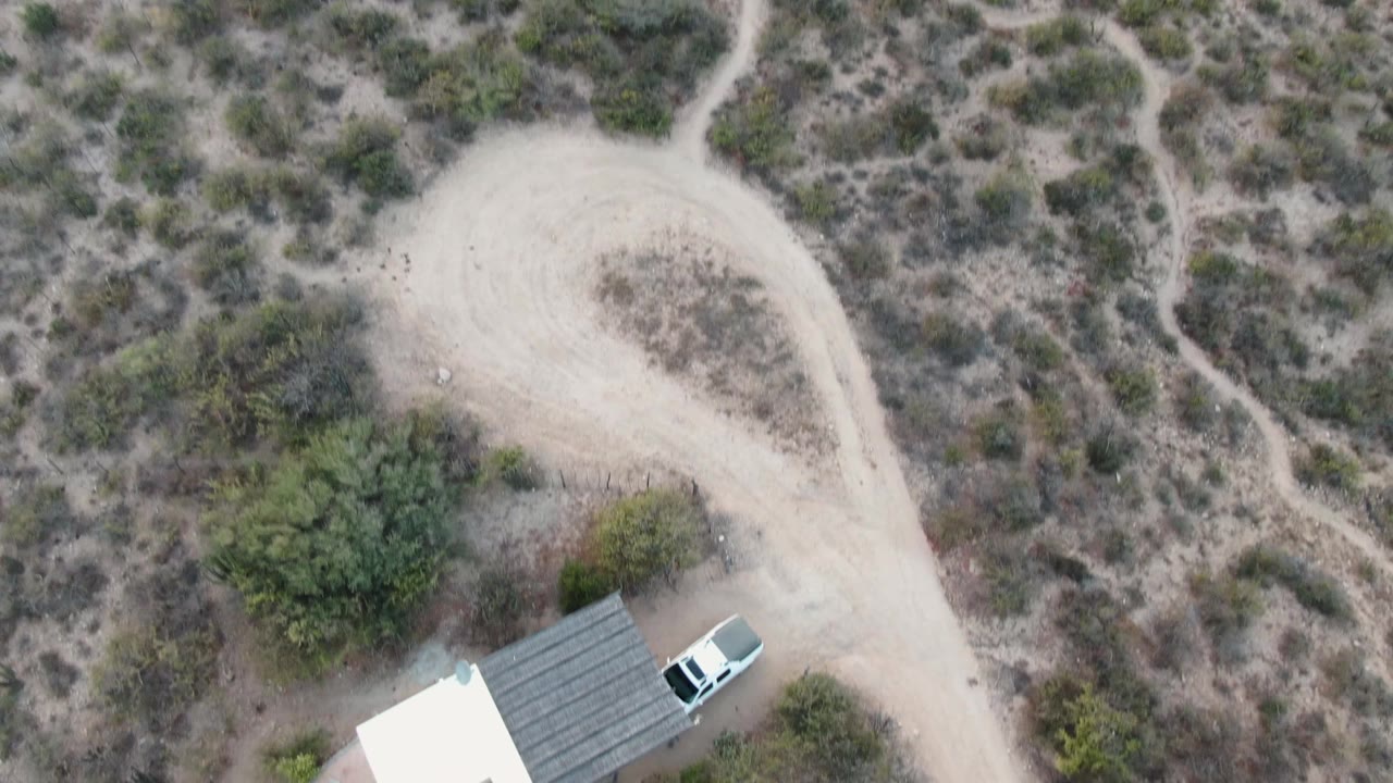Aerial view of a small structure, white vehicle, pool edge, and dirt road.