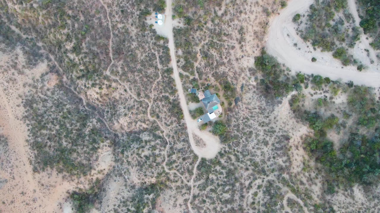 Overhead frame of Cabo Pulmo desert terrain with a pale dirt road beside a small light-roofed structure, scattered scrub, and no visible people.