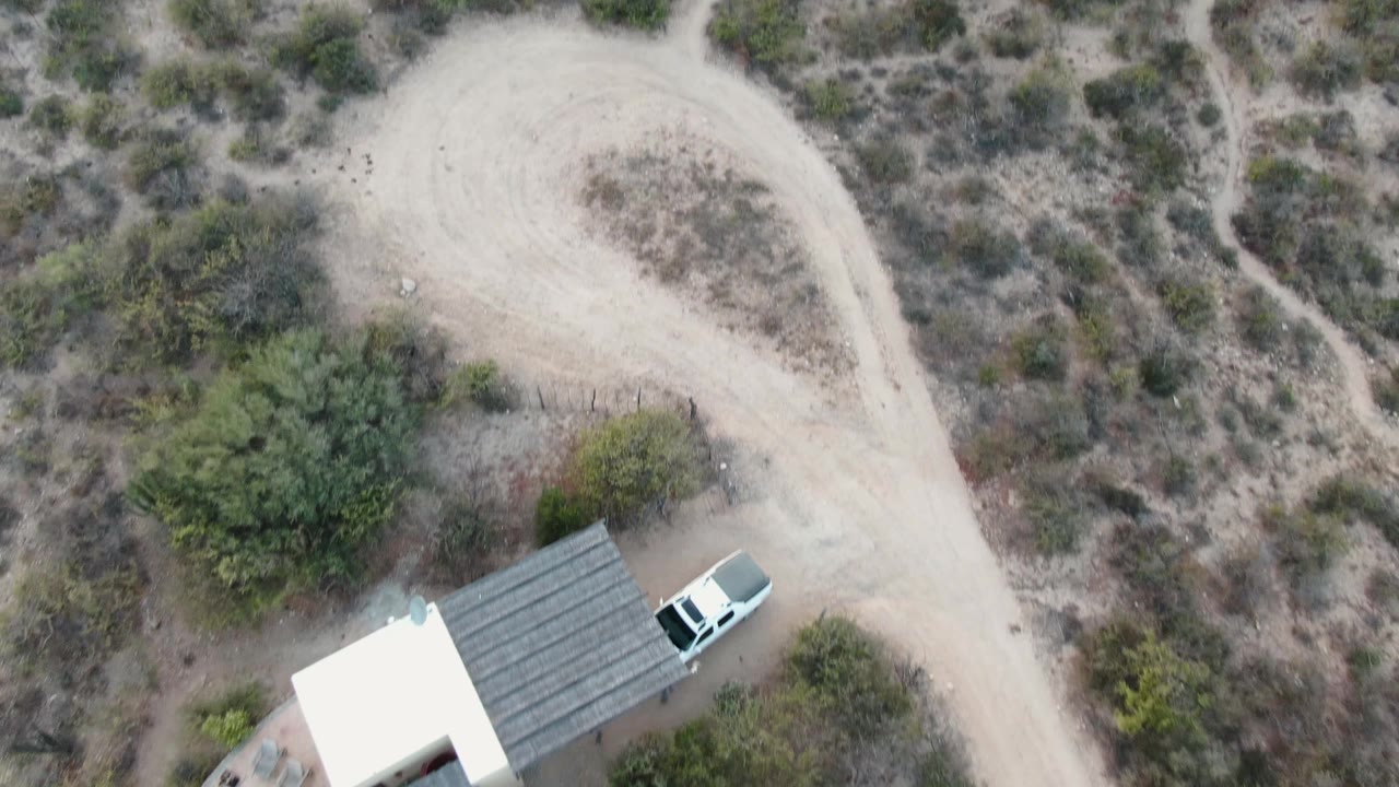 Overhead view of a small roofed shelter, white car, dirt road, and dry scrub.
