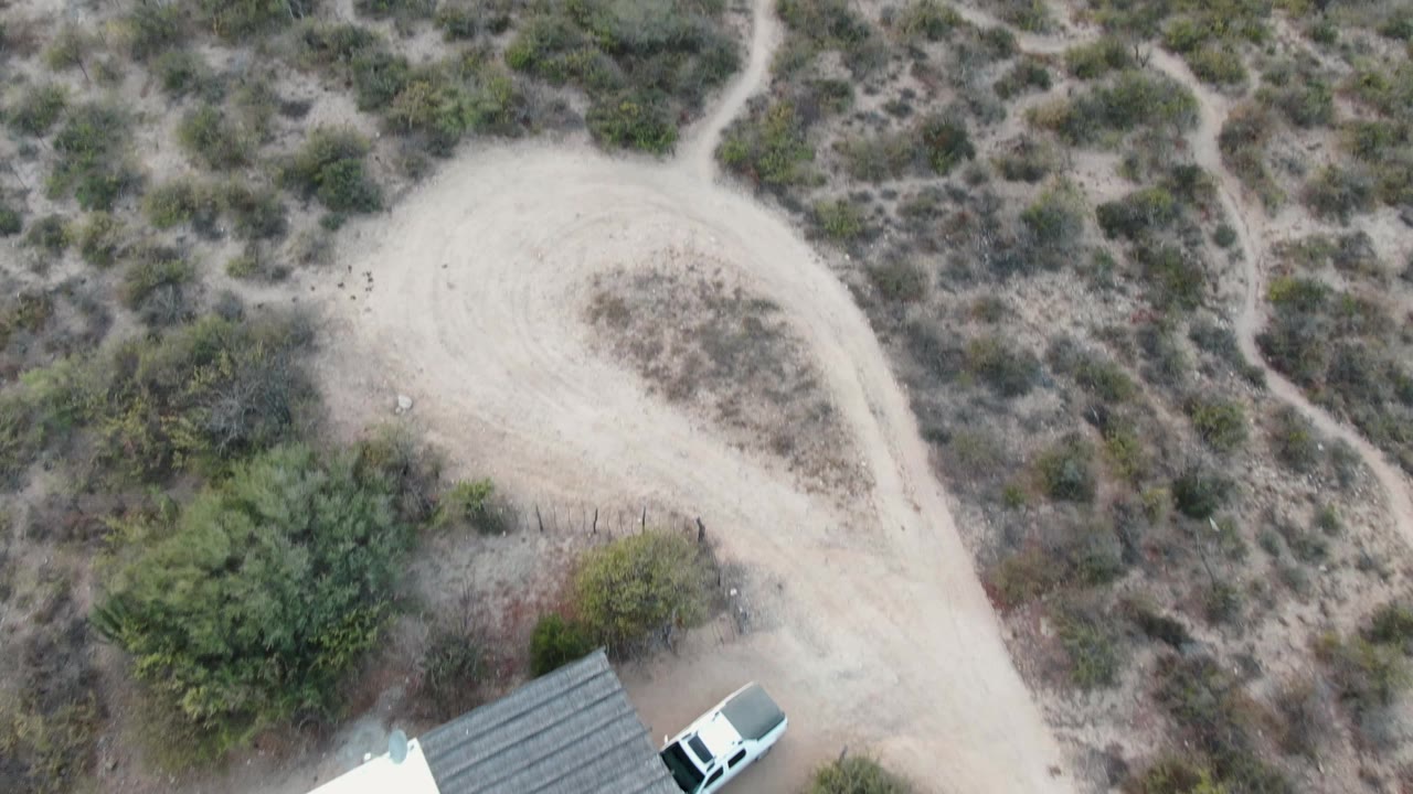A Cabo Pulmo aerial frame with the roof edge and white car partly visible at the bottom, a curved dirt track above, scrubby vegetation, and no visible people.
