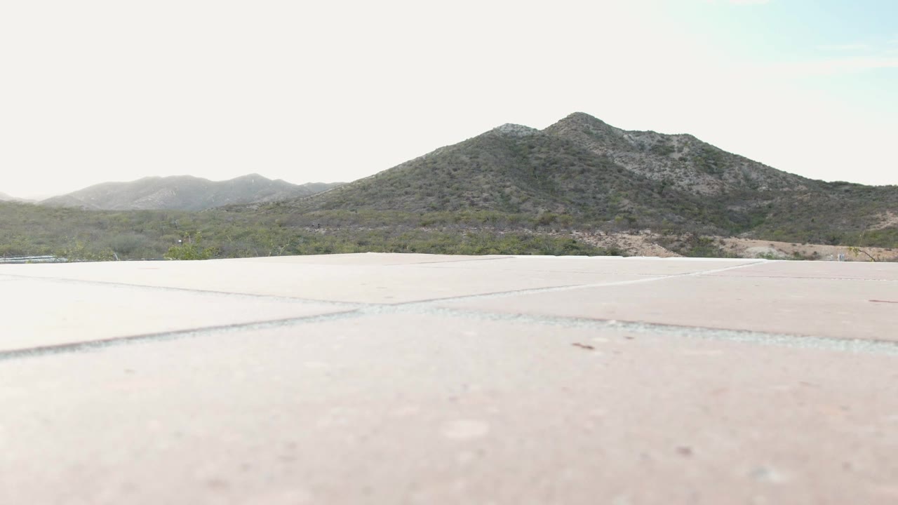At one second, the Cabo Pulmo frame holds on the pale paved foreground, low brush across the midground, a dark mountain ridge, and no visible people.
