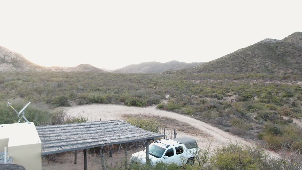 At 0:00 in Cabo Pulmo, Baja California Sur, a pale building, slatted carport, and white truck sit below scrub-covered hills with no visible people.