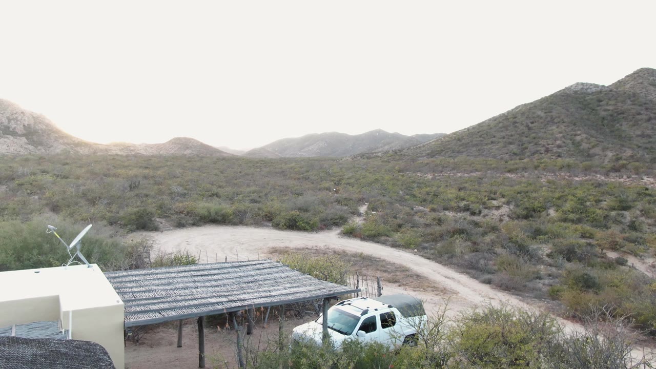 At 0:01, the white truck is more centered beside the slatted carport, with a winding dirt road and Cabo Pulmo desert brush behind it and no visible people.