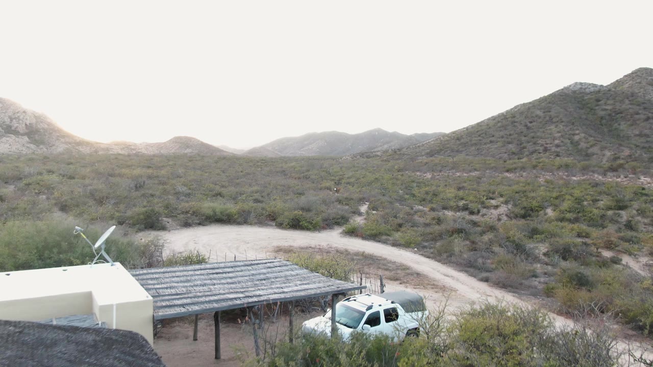 At 0:02, the elevated view keeps the carport and white truck low in frame while the Cabo Pulmo dirt road curves through scrub toward distant hills, with no visible people.