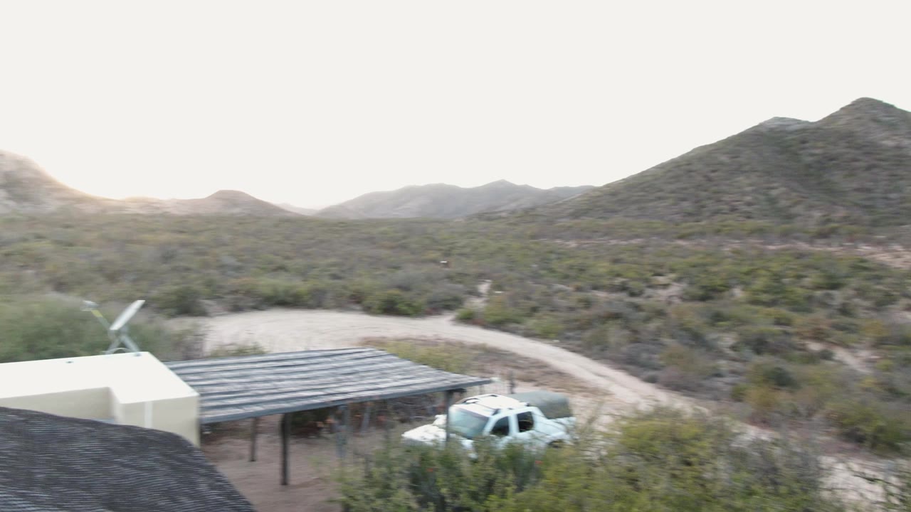 Pale building, carport roof, dirt road, and scrubby desert hills.