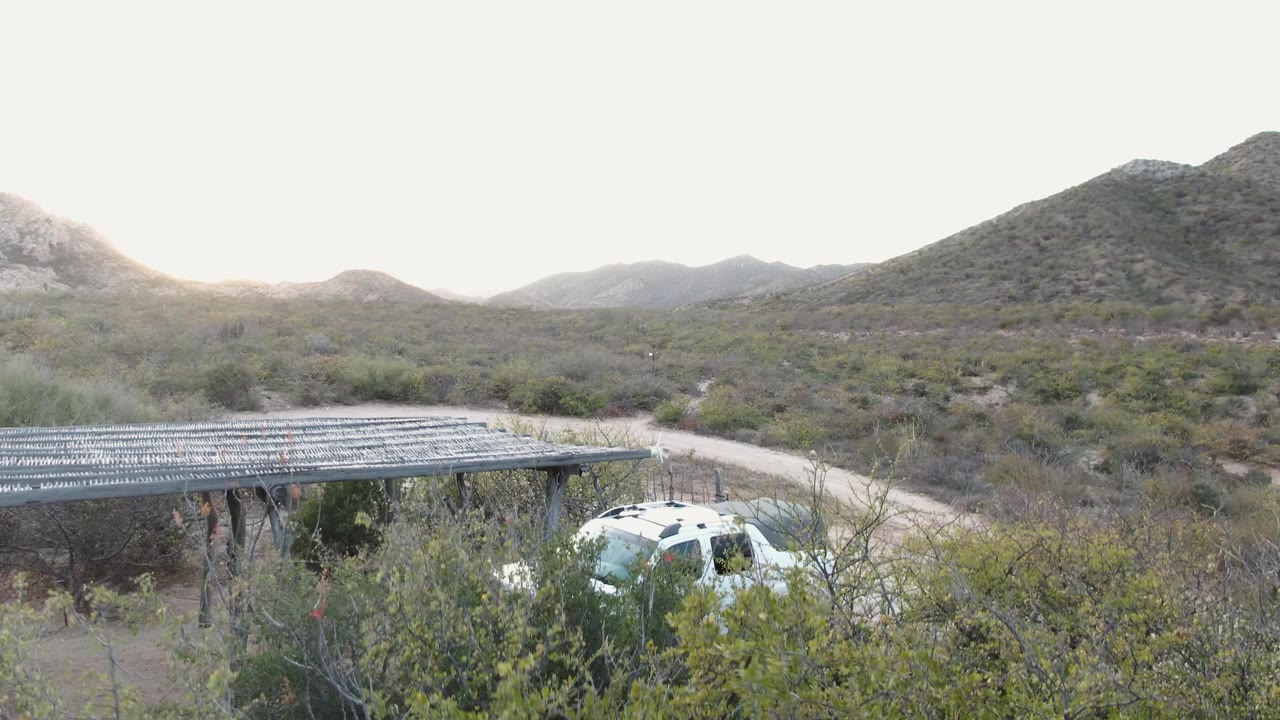 A white SUV sits partly hidden by foreground brush beside a wooden shelter in Cabo Pulmo, Baja California Sur, with low desert hills behind and no visible people.